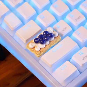 Może przedstawiać: Miniaturowe ciasto z jagodami i kremem leży na białej klawiaturze. Ciasto ma złoty brązowy kolor, z warstwą kremu i zwieńczone kilkoma ciemnoniebieskimi jagodami. Klawisze klawiatury są białe z niebieskim podświetleniem.