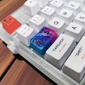May include: Close-up of a mechanical keyboard with custom keycaps. The keycaps are white, with a few accent keys in red and a colorful, artistic design. The "CAPSLOCK" and "SHIFT" keys are visible, along with other keys.