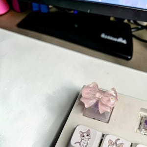 May include: A light pink, bow-shaped keycap on a white keyboard. The keycap is translucent, with a small yellow accent in the center. The keyboard also features cat-themed keycaps.