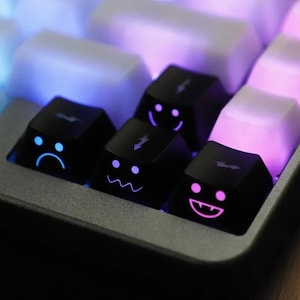 May include: Close-up of a mechanical keyboard with black keycaps featuring emoji faces. The keycaps have blue and pink backlighting. The emoji faces include a sad face, a lightning bolt, a wavy mouth, and a smiling face.