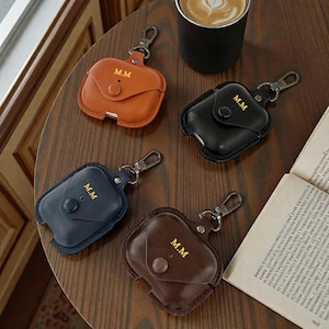 May include: Four leather AirPod cases in shades of brown, black, and blue, each with a silver clasp and the initials "M.M." embossed in gold. The cases are arranged on a wooden surface, with a cup of coffee and a book in the background.