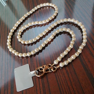 Puede incluir: Un cordón para teléfono con cuentas de color crema con detalles dorados y un cierre dorado. El cordón presenta cuentas de imitación perla alternadas y cuentas más pequeñas y brillantes. Se adjunta un soporte para teléfono transparente y rectangular.