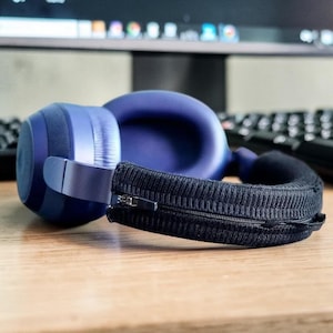 Può includere: Cuffie blu over-ear con una copertura nera con cerniera. Le cuffie hanno un archetto e padiglioni imbottiti. La copertura è realizzata in tessuto strutturato. Le cuffie sono appoggiate su una superficie di legno.
