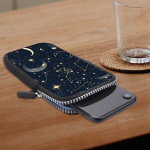 Pode incluir: Um smartphone preto parcialmente dentro de uma capa com zíper. A capa apresenta um design celeste azul escuro com estrelas douradas, constelações e luas crescentes. O zíper é prateado e a capa está sobre uma superfície de madeira.