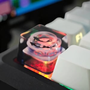 May include: Close-up of a custom keyboard keycap. The keycap is transparent with a design inside, and it is lit with red and orange light. Other white keycaps are visible in the background.