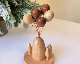 May include: A wooden decorative piece featuring a cluster of wooden balloons in varying shades of brown and beige. The balloons are attached to a wooden vase-like base with small wooden tree-like accents. The piece is set on a round wooden base.