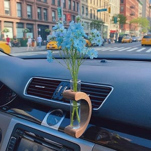 May include: A car air vent vase with a wooden holder, holding a small glass tube with blue flowers. The vase is set against a dark dashboard, with a city street in the background.