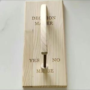 May include: A wooden decision maker with the words "DECISION MAKER", "YES", "NO", and "MAYBE" engraved in a dark brown font. A light-colored wooden handle is attached to a dark metal rod, which is connected to the wooden base.