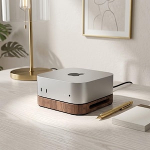 May include: A silver Apple Mac Mini computer sits atop a dark brown wooden stand. The device features a square shape with rounded corners, a black Apple logo on the top surface, and a small circular indicator light on the front panel.
