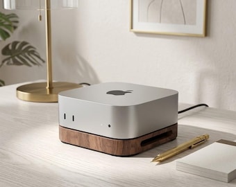 Może przedstawiać: Srebrny komputer Apple Mac Mini stoi na ciemnobrązowej drewnianej podstawce. Urządzenie ma kwadratowy kształt z zaokrąglonymi rogami, czarne logo Apple na górnej powierzchni oraz małą okrągłą lampkę kontrolną na przednim panelu.