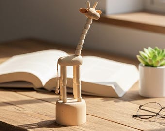May include: A wooden giraffe figurine with a long, segmented neck and small ears. The giraffe stands on a round base and has jointed legs. The figurine is light brown and sits on a wooden surface near an open book and a small potted plant.