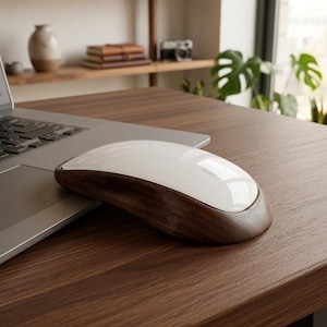 May include: A computer mouse featuring a smooth, glossy white top surface and a dark brown wood grain base. The mouse is positioned on a wooden desk surface next to a silver laptop, showcasing a contrast between the white finish and wood texture.