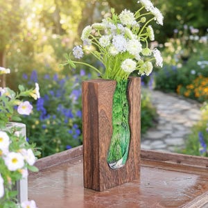 May include: A wooden vase with a rectangular shape and a green, organic-shaped glass insert. The vase holds a bouquet of white and light purple flowers. The wood has a dark brown finish.