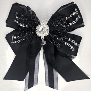 May include: A black bow with a decorative silver brooch at the center. The bow features layers of black ribbon, lace, and sheer fabric. Silver-colored beaded accents and safety pins add detail. A dangling crystal hangs from the brooch.