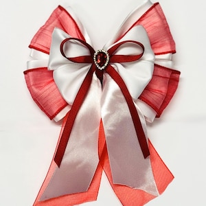 May include: A decorative bow featuring layers of white and red fabric. The bow has a central white satin section with a red ribbon and a sparkling jewel accent. The edges are trimmed with a sheer red material.