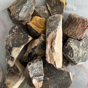 Op de afbeelding: Een verzameling ruwe, natuurlijke vuurstenen in verschillende maten en vormen. De stenen vertonen een reeks kleuren, waaronder donkergrijs, bruin, beige en hints van oranje. Sommige stukken hebben een ruw, getextureerd oppervlak.