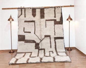 May include: A large, rectangular rug with a geometric pattern in shades of brown and cream. The rug is hanging on a wooden rod against a white wall. Two copper-colored floor lamps flank the rug.