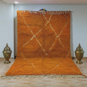 May include: An orange Moroccan rug with a diamond pattern in white. The rug has a shaggy texture and a fringe along the bottom edge. Two decorative gold-colored lamps flank the rug.
