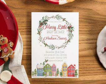 May include: A white baby shower invitation card featuring the text "Merry Little Baby Shower" and the name "Madison Suarez". The card is decorated with a wreath and a watercolor illustration of colorful houses and trees. The invitation is placed on a wooden surface.
