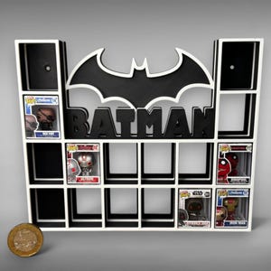 May include: A black and white wall-mounted display shelf featuring a large Batman logo and the word BATMAN in bold lettering. The unit has multiple square compartments, some containing small collectible figures. A gold coin sits to the left.
