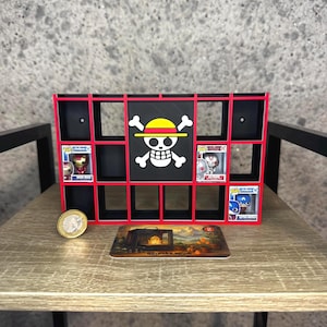 May include: A black and red display shelf with multiple compartments, featuring a pirate flag design in the centre. The shelf holds several collectible figurines. A coin and a decorative card are in front of the shelf.