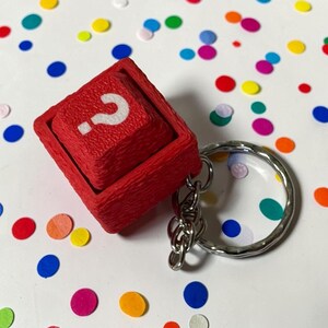 May include: A red, cube-shaped keyring featuring a raised, square button on its top surface. The button is textured and displays a white question mark symbol. A silver-coloured metal split ring is attached to the cube by a short metal chain.