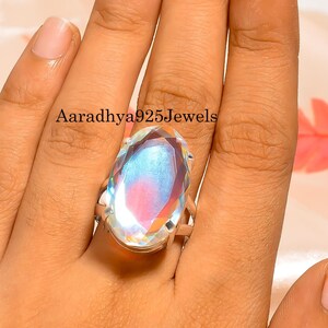 May include: A large, oval-cut gemstone ring with a silver band. The gemstone displays a rainbow of colors, including blue, pink, and yellow. The ring is shown on a hand, with the text "Aaradhya925Jewels" visible.