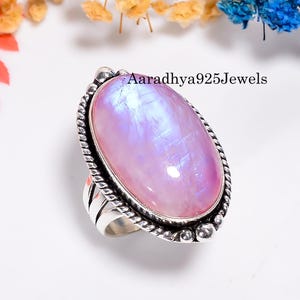 May include: A silver ring featuring a large, oval-shaped moonstone with a pearlescent, pinkish-white hue. The stone is set in a detailed, rope-like bezel. The ring is from Aaradhya925Jewels.