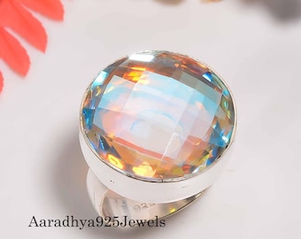 Könnte beinhalten: Ein Silberring mit einem großen, facettierten, irisierenden Edelstein. Der Edelstein zeigt eine Reihe von Farben, darunter Gelb, Orange und Blau. Der Ring ist vor einem weißen Hintergrund mit dekorativen Blumen und Blättern platziert.