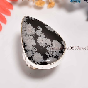 Puede incluir: Un anillo de plata con una gran piedra preciosa negra en forma de lágrima con un patrón de copo de nieve. La piedra está engastada en una banda de plata pulida. El texto "Aaradhya925Jewels" es visible en la piedra.