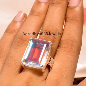May include: A large, rectangular gemstone ring with a silver band. The gemstone has a vibrant, iridescent colour with red, orange, and blue hues. The ring is worn on a finger, and the text "Aaradhya925Jewels" is visible.