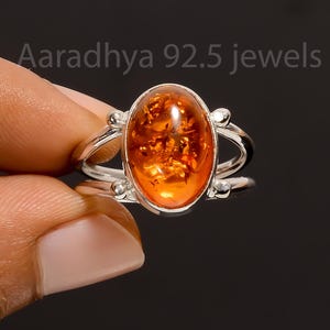 May include: A silver ring with an oval amber gemstone. The amber is a warm orange hue, set in a silver band with decorative details. The ring is held by a hand against a dark backdrop. The image includes the text "Aaradhya 92.5 jewels."