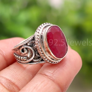 May include: A silver ring with a large, oval-shaped, deep red gemstone. The ring has an ornate, detailed design with scrollwork and a textured band. The text "92.5 jewels" is visible on the ring.