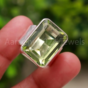 May include: A silver ring with a large, rectangular, pale green gemstone. The gemstone is cut with facets, catching the light. The ring is held in a hand, with the text "Aarav 925 Jewels" visible.