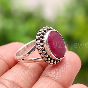 Könnte beinhalten: Ein Silberring mit einem großen, ovalen Rubin-Edelstein. Der Rubin hat eine tiefrote Farbe und ist von einem perlenbesetzten Rand umgeben. Der Ring hat ein geteiltes und strukturiertes Band. Der Text "925 jewels" ist sichtbar.