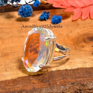 May include: A silver ring with a large, faceted, oval-shaped gemstone. The gemstone has a rainbow iridescence and is set in a split-shank band. The text "Aaradhya925Jewels" is visible in the image.