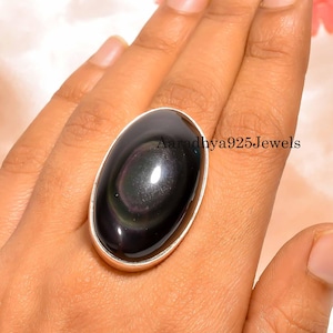 Könnte beinhalten: Ein großer, ovaler Ring mit einem polierten schwarzen Obsidianstein mit schillernden Wirbeln. Der Stein ist in einem silberfarbenen Metallband gefasst. Der Ring wird auf einer Hand gezeigt.