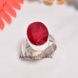 May include: A silver ring featuring a large, oval-cut, vibrant red gemstone. The ring has a wide band and a simple, elegant design. The text "Aaradhya925Jewels" is visible on the ring.