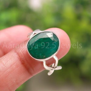 May include: A silver ring featuring a large, oval-cut emerald gemstone. The emerald is a deep green color and is set in a silver band with a floral design. The text "Aaradhya 92.5 jewels" is visible.