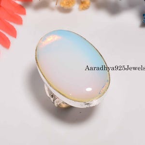 Può includere: Un anello d'argento con una gemma opalescente ovale. La pietra ha un colore bianco perlaceo e azzurro. L'anello è incastonato in una fascia d'argento. Il testo "Aaradhya925Jewels" è visibile.