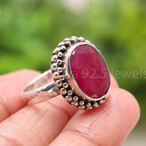Könnte beinhalten: Ein Silberring mit einem großen, ovalen Rubin-Edelstein. Der Rubin hat eine tiefrote Farbe und ist von einem perlenbesetzten Rand umgeben. Der Ring wird von einer Hand gehalten, mit dem Text "a 92.5 jewels" sichtbar.