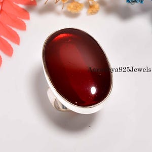 Pode incluir: Anel de prata com uma grande pedra preciosa ovalada de cor vermelha escura. A pedra preciosa está engastada em uma moldura. O texto "Aaranya925Jewels" é visível no lado direito do anel.