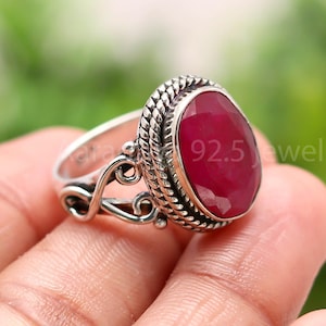 Pode incluir: Um anel de prata com uma grande pedra preciosa de rubi em forma oval. O anel apresenta uma moldura trançada e filigrana decorativa na banda. O texto "92.5 jewels" é visível no anel.