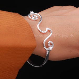 Peut inclure: Un bracelet manchette ouvert de couleur argentée avec un design abstrait et fluide. Le bracelet présente une forme ondulée et organique avec des volutes arrondies aux extrémités. Le bijou est porté sur un poignet, sur fond noir.