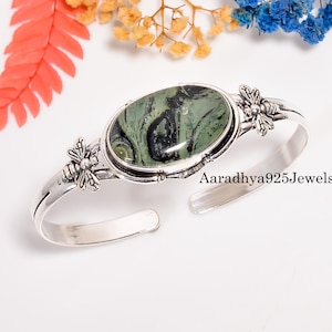 May include: Silver cuff bracelet featuring an oval green and black stone. The bracelet has bee-shaped accents on each side of the stone and the text "Aaradhya925Jewels". A stylish piece of jewelry.