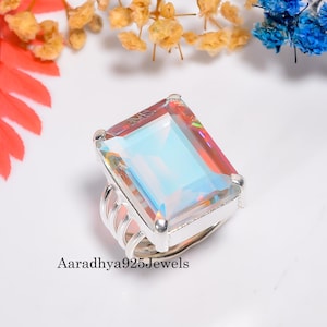 May include: A rectangular iridescent gemstone ring with a silver band. The gemstone reflects multiple colors, including blue, pink, and yellow. The ring is set against a white background with colorful floral accents. The text "Aaradhya925Jewels" is visible.