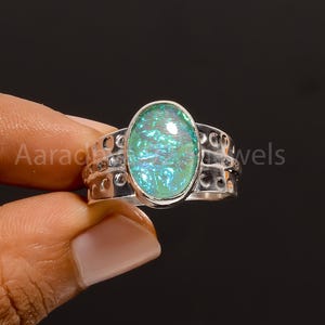 Könnte beinhalten: Ein silberner Ring mit einem ovalen, irisierenden, hellgrünen Edelstein. Das Ringband hat ein dekoratives Muster mit kleinen kreisförmigen Aussparungen. Der Ring wird von den Fingern einer Person vor dunklem Hintergrund gehalten. Der Text "Aaradhya Jewels" ist sichtbar.