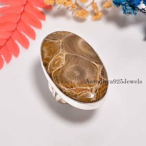 Pode incluir: Anel oval com banda prateada, apresentando uma pedra estampada em tons de castanho e bege. O anel está sobre um fundo branco com detalhes florais coloridos. O texto "Aaradhya925Jewels" é visível.