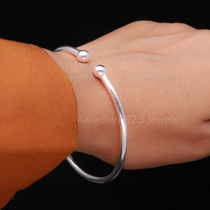 May include: A silver cuff bracelet with rounded ball ends. The open design allows for easy wear. The bracelet is shown on a person's wrist against a black background. The text "Aaradhy 92.5 jewels" is visible.