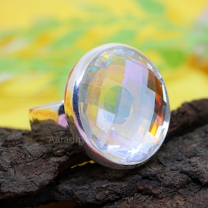 May include: A silver ring featuring a large, faceted, clear gemstone. The gemstone reflects a rainbow of colors. The ring is set on a dark, textured surface with a yellow background.